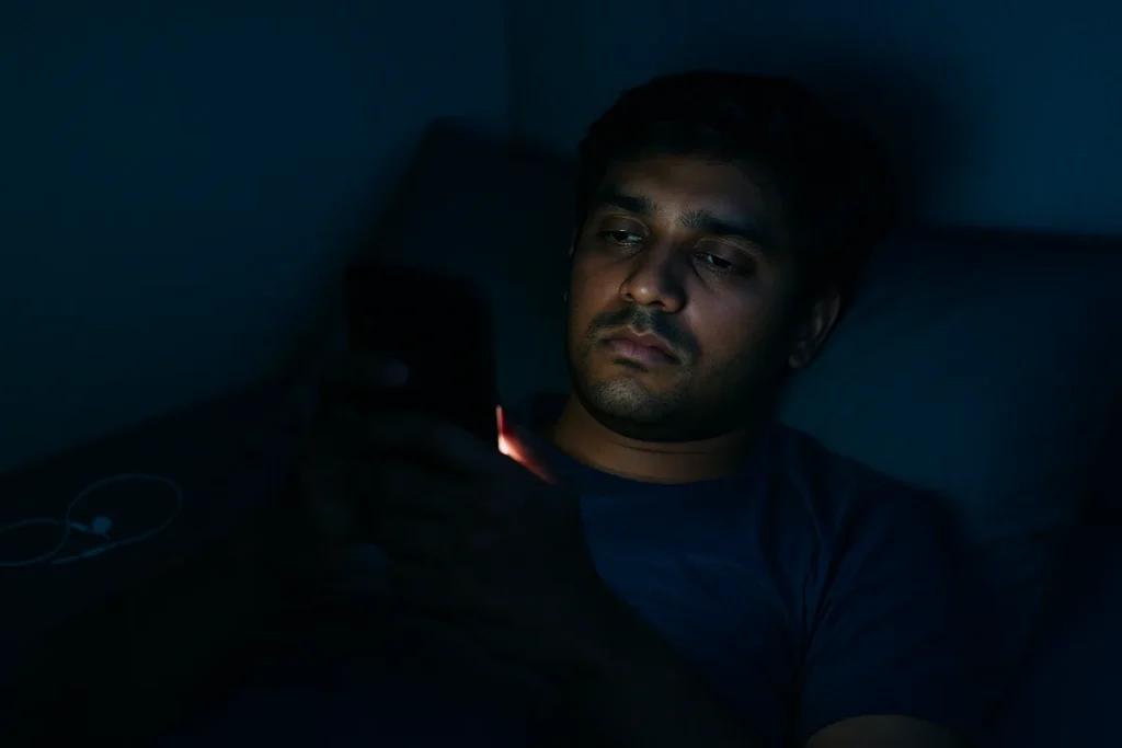 రాత్రి మంచంపై పడుకుని మొబైల్ చూస్తూ మానసికంగా అలసిపోయిన యువకుడు. స్క్రీన్ కాంతి అతని ముఖంపై మసకబారగా పడుతోంది.