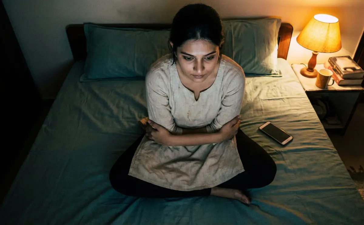 రాత్రి గదిలో మంచంపై మోకాళ్లపై కూర్చుని, చేతులు మడిచుకుని, లోపల దాచిన భయాలతో ఆలోచనల్లో మునిగిన యువతి