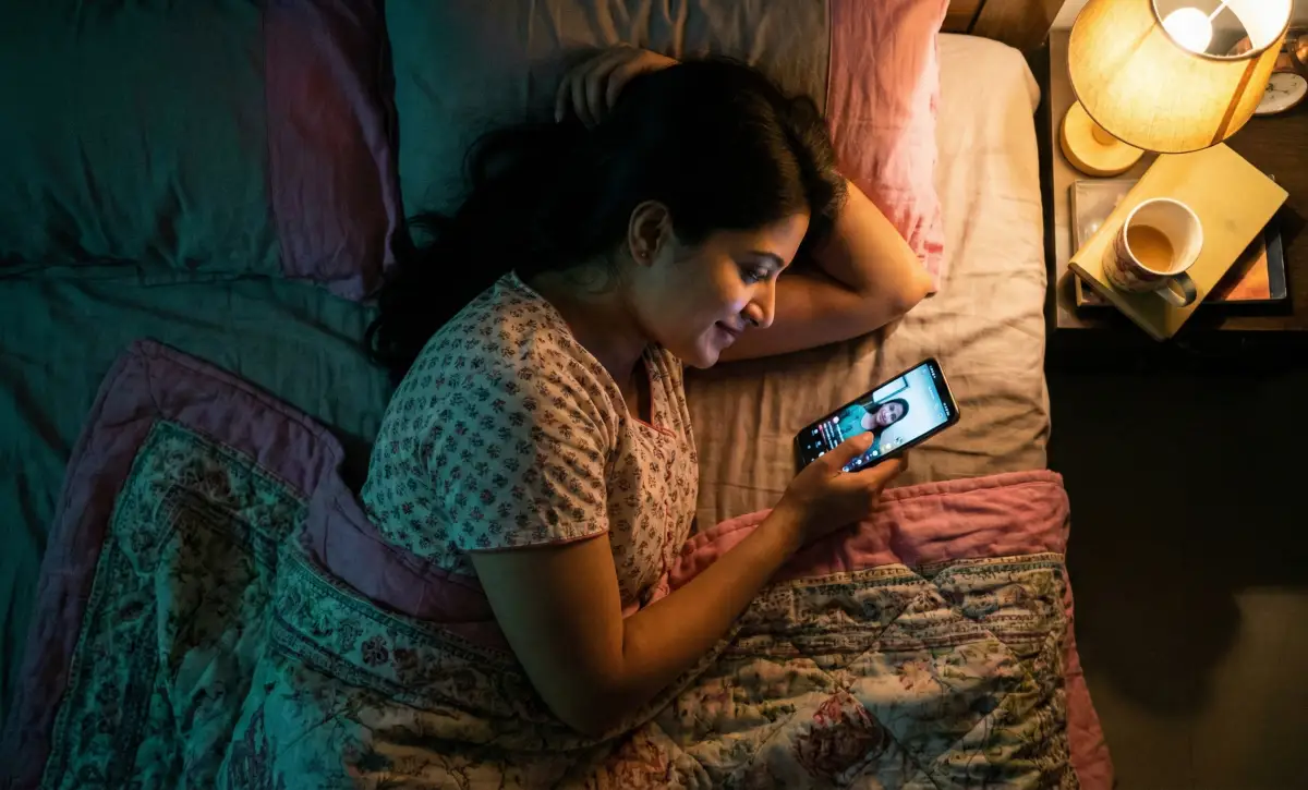 రాత్రి మంచంపై పక్కగా పడుకుని, మొబైల్ స్క్రీన్‌లో వీడియో కాల్ చూస్తూ మృదువుగా నవ్వుతున్న యువతి